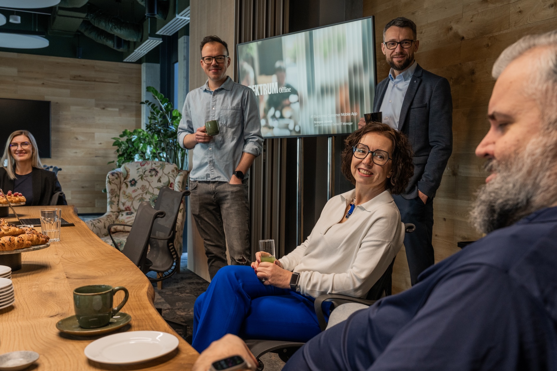 Grupa uczestników wydarzenia Spektrum Offline uśmiecha się i rozmawia przy stole. Dwie osoby stoją przy ekranie z prezentacją, trzy siedzą z napojami w rękach. W tle widoczny ekran z tytułem prezentacji oraz jasne, nowoczesne biuro z roślinami i fotelami.