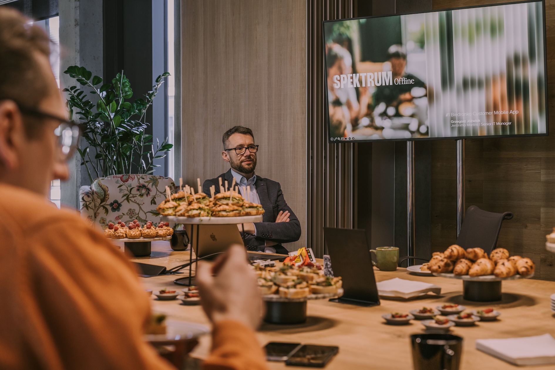 Mężczyzna w garniturze siedzi przy stole podczas spotkania. Na ekranie za nim widoczny jest slajd tytułowy prezentacji Spektrum Offline: ‘Biedronka Customer Mobile App’. Przed uczestnikami znajdują się tace z kanapkami i przekąskami. Na pierwszym planie rozmyty profil jednej z osób oraz talerz z jedzeniem.