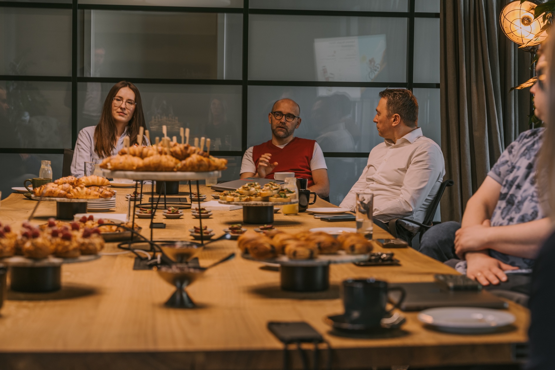 Uczestnicy spotkania Spektrum Offline siedzą przy stole zastawionym przekąskami i rozmawiają. W centrum kadru widoczna jest osoba zabierająca głos, wokół niej pozostali słuchacze. W tle – przeszklona sala i prezentacja wyświetlana na ekranie.