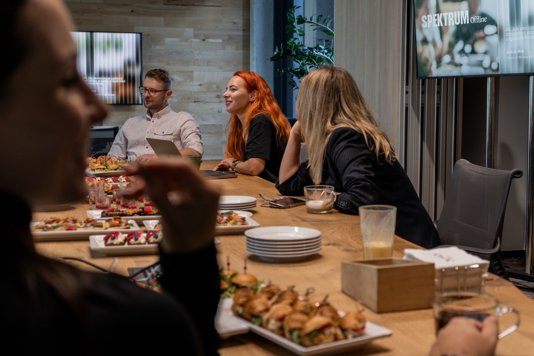 Zdjęcie z wydarzenia Spektrum Offline #4 w biurze SYZYGY Warsaw. W centrum kadru widać Jakuba Raka oraz uczestniczki rozmowy, siedzących przy stole zastawionym kanapkami, croissantami i deserami. Na pierwszym planie rozmazana sylwetka osoby sięgającej po przekąskę. W tle ekran z logotypem Spektrum Offline.