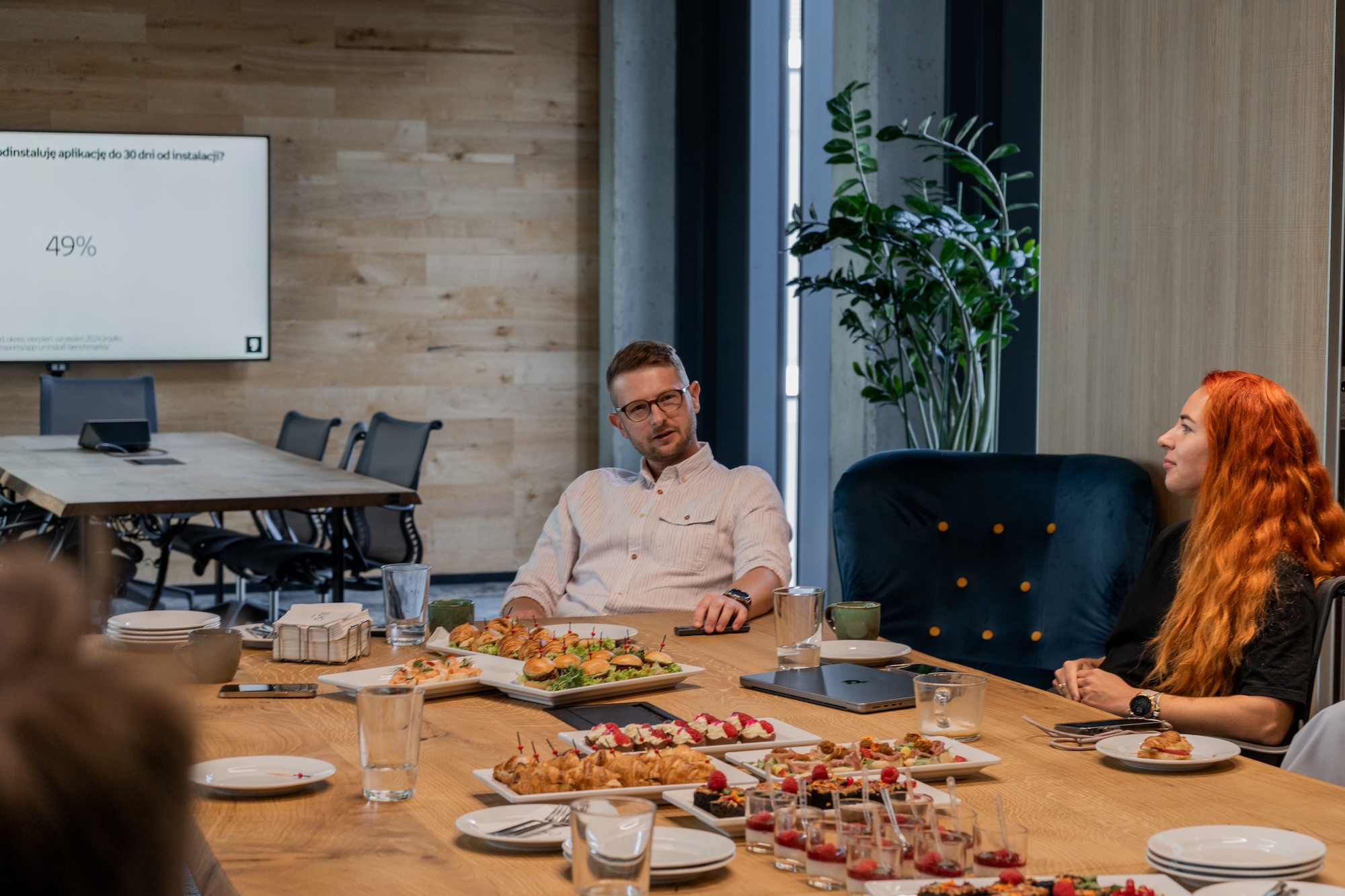 Zdjęcie z wydarzenia Spektrum Offline #4 w biurze SYZYGY Warsaw. Jakub Rak, Head of Mobile w Empik Group, prowadzi rozmowę z jedną z uczestniczek o lojalności użytkowników aplikacji. Na stole widać poczęstunek: kanapki, croissanty i desery, a w tle ekran ze slajdem pokazującym wynik „49%”.