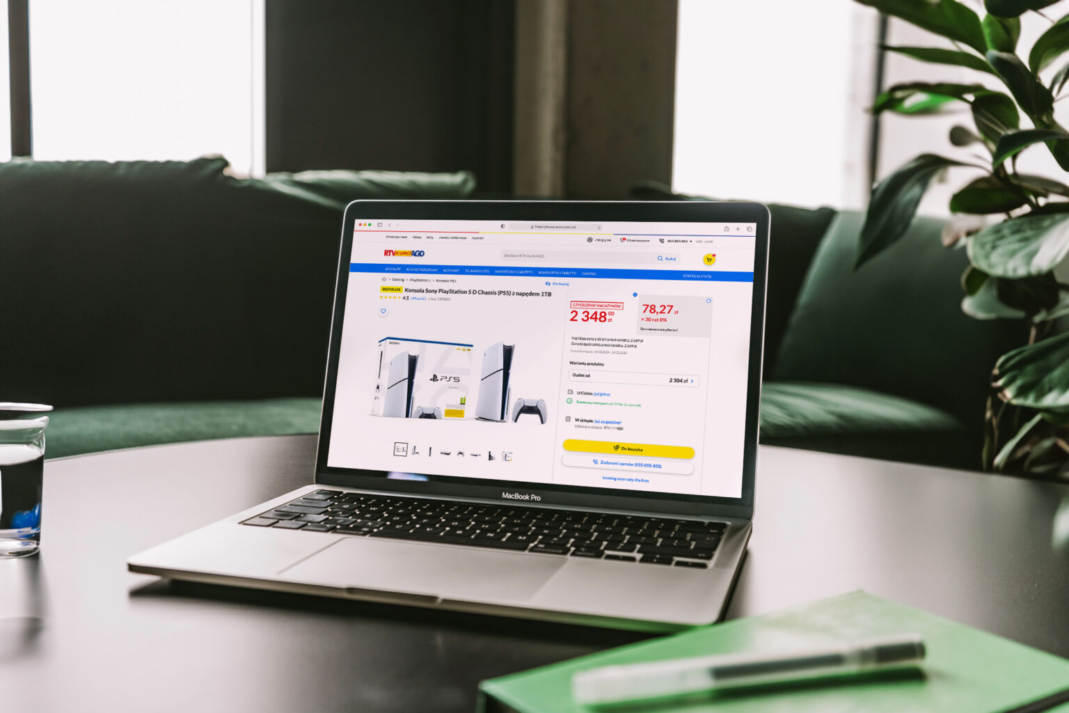 Laptop MacBook Pro na stole w nowoczesnym wnętrzu, z otwartą stroną internetową RTV EURO AGD, przedstawiającą konsolę Sony PlayStation 5 z napędem 1TB w ofercie sprzedaży. Na ekranie widoczne są szczegóły produktu, cena oraz opcje zakupu. Obok laptopa znajduje się szklanka z wodą i zielony notes. W tle zielona sofa oraz rośliny.