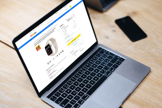 RTV EURO AGD - platforma e-commerce - Laptop z otwartą stroną sklepu internetowego RTV EURO AGD, prezentującą smartwatch Apple Watch SE 2 z białym paskiem. Na ekranie widoczne są szczegóły produktu, cena, opcje zakupu oraz przycisk „Do koszyka”. Laptop stoi na drewnianym stole, w tle znajduje się czarny smartfon.