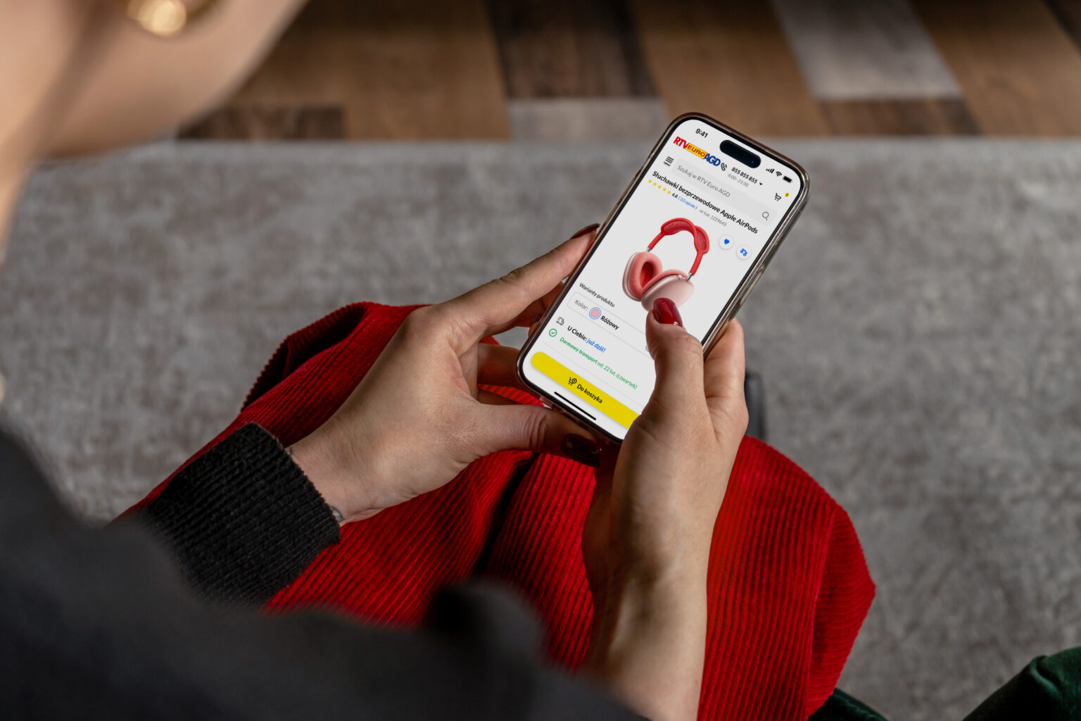 Mobilna wersja sklepu RTV EURO AGD - platformy e-commerce - Osoba trzymająca w dłoniach smartfon z otwartą stroną sklepu internetowego RTV EURO AGD, prezentującą słuchawki bezprzewodowe Apple AirPods Max w kolorze różowym. Na ekranie widoczne są szczegóły produktu, opcje zakupu oraz przycisk „Do koszyka”. W tle widać fragment czerwonej spódnicy i drewnianą podłogę.